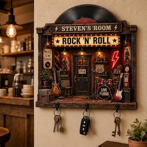 Könnte beinhalten: Ein wandmontierter Schlüsselhalter mit einem Miniatur-Rock-and-Roll-Zimmer-Diorama. Das Schild zeigt STEVEN'S ROOM und ROCK 'N' ROLL. Es enthält ein Schlagzeug, Gitarren, Verstärker und ein Route-66-Schild. Drei schwarze Metallhaken halten unten Schlüssel.