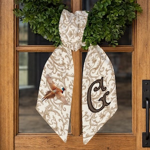 May include: A decorative door bow with a pheasant illustration and the letter "G". The bow is cream-colored with a brown floral pattern. The pheasant is depicted in flight with brown, red, and blue feathers. The bow is hanging on a wooden door.