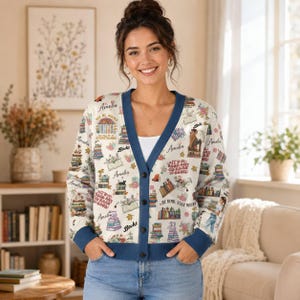 May include: A white long-sleeve cardigan with a blue trim and a repeating pattern of colourful books, floral accents, and the text "Amelia," "Books," and "It's a good day to read a book." The cardigan features a V-neckline and a button-down front closure.