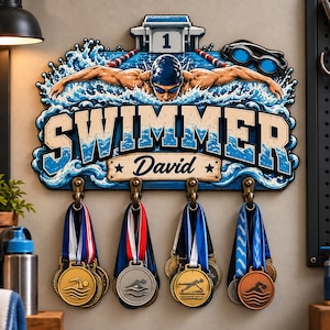 Könnte beinhalten: Ein wandmontierter Medaillenhalter mit einer Grafik eines Schwimmers in einem Becken und dem aufgedruckten Text "SWIMMER" und "David". Das Schild verfügt über vier bronzefarbene Metallhaken, an denen jeweils mehrere Sportmedaillen hängen.