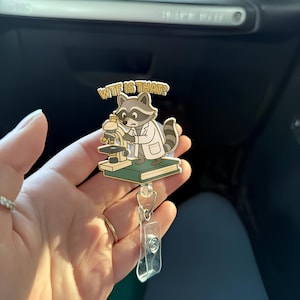 May include: A badge reel featuring a cartoon raccoon in a lab coat looking through a microscope. The raccoon is standing on a green book, with the text "WTF IS THAT?" above it. The reel has a clear plastic clip.