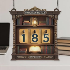 Może przedstawiać: Dekoracyjna drewniana półka na książki z napisem "BOOKS READ THIS YEAR" i "SOPHIA'S LIBRARY". Półka wyświetla numer "185" na trzech podświetlanych panelach. Zawiera miniaturowe książki, latarnię i jest zawieszona na łańcuchu.