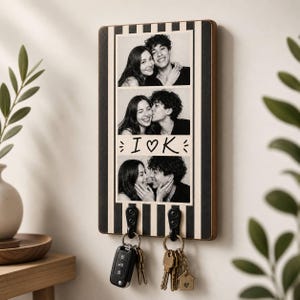 May include: A rectangular wooden wall-mounted key holder featuring a black and cream vertical striped pattern. The center displays three black and white photographs of a couple, with the text I heart K printed between the images. Two black metal hooks hold sets of keys.