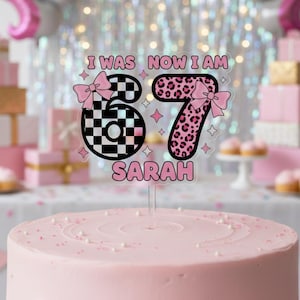 Puede incluir: Una tarta glaseada rosa coronada con un adorno transparente para tarta. El adorno dice "I WAS NOW I AM 67 SARAH" con un 6 a cuadros y un 7 con estampado de leopardo rosa, ambos adornados con lazos rosas. También se incluyen estrellas rosas y un lazo rosa.