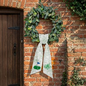 May include: A green eucalyptus wreath hangs on a brick wall, adorned with a white scarf featuring a golf course design and the letter "M". The golf course design includes a green putting green, a blue flag, and a golf ball.