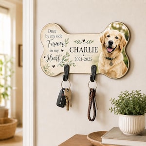 May include: A bone-shaped wall-mounted key holder featuring a photo of a golden retriever, the text "Once by my side Forever in my Heart", the name "CHARLIE", and the years "2021-2025". It includes two black metal hooks holding sets of keys.