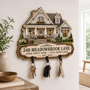 May include: A wall-mounted wooden key holder shaped like a house with a gray roof and white siding. The sign features the text 248 MEADOWBROOK LANE, HOME SWEET HOME, LOVE GROWS HERE, and EST. 2026. Three black metal hooks hold various keys and a fob.