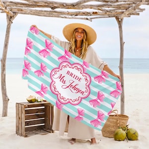 May include: A beach towel with a striped pattern in white and light blue, adorned with pink bows. The towel features the text "Bride Mrs Johnson" in a decorative frame. The towel is held up on a beach.