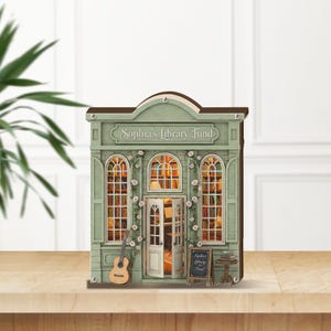 May include: A miniature, light green library model with the text "Sophia's Library Fund" above the windows. The model features a guitar, open doors, and a small chalkboard sign. The windows are lit, suggesting a cozy interior.