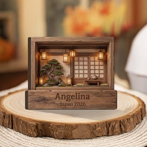 Pode incluir: Diorama em caixa de sombra de madeira com uma cena de jardim japonês em miniatura. Apresenta um bonsai, lanternas e um serviço de chá. A base de madeira é gravada com "Angelina" e "Japan 2026". Exibido em uma fatia de madeira.