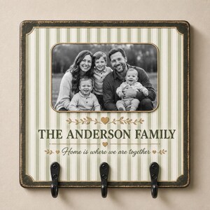 May include: A square wall-mounted key holder with a distressed black border and a cream and sage green striped background. It features a central black and white family photo, the text THE ANDERSON FAMILY, and Home is where we are together.