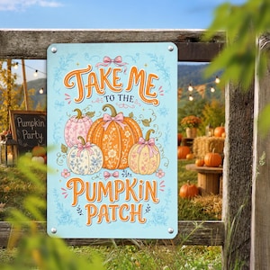 Puede incluir: Un letrero rectangular de metal azul claro con el texto naranja "TAKE ME TO THE PUMPKIN PATCH" y una ilustración de tres calabazas en tonos naranja, rosa y blanco, decoradas con patrones florales y pequeños lazos rosas.