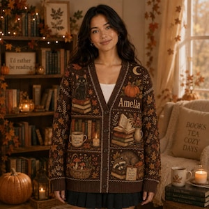 May include: A brown V-neck cardigan featuring a knit pattern of autumn leaves, a black cat on books, a pumpkin, a mug, a candle, a basket of mushrooms, a raccoon, and the name Amelia. The design includes open books and the text Books are my happy place.