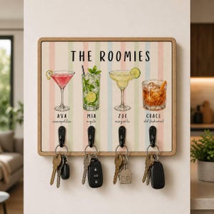 May include: A rectangular wooden wall-mounted key holder featuring a pastel striped background and the text THE ROOMIES. It displays four cocktail illustrations labeled Ava cosmopolitan, Mia mojito, Zoe margarita, and Grace old fashioned, with four black metal hooks holding various keys.