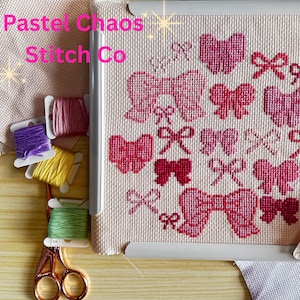 May include: A cross-stitch design with pink bows and hearts on a light beige fabric, framed in white. The text "Pastel Chaos Stitch Co" is visible. Spools of colourful embroidery floss and scissors are also present.