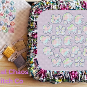 May include: A cross-stitch project in progress, featuring a lavender-colored fabric with a design of pastel-colored hearts, stars, and butterflies. The project is framed by a colorful, patterned fabric. Three spools of DMC embroidery floss are visible.