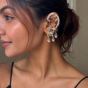 May include: An ornate ear cuff featuring a curved gold-tone frame adorned with small white pearls and a vibrant array of multicoloured round gemstones. The design includes three dangling teardrop-shaped clear crystals at the base of the earring.