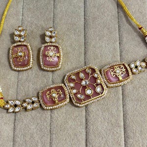 Könnte beinhalten: Ein goldfarbenes Halsketten- und Ohrring-Set. Die Halskette hat rosa, quadratische Anhänger mit aufwendigen Golddesigns und klaren Steinakzenten. Die Ohrringe spiegeln das Anhängerdesign wider. Das Set befindet sich auf einem neutralen Hintergrund.