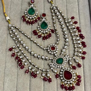Peut inclure: Un ensemble de bijoux orné comprenant un collier à plusieurs rangs et des boucles d'oreilles assorties. Le collier et les boucles d'oreilles sont ornés de pierres précieuses transparentes, de pierres vert émeraude et rouge rubis, et de petites perles. L'ensemble est présenté sur une surface de couleur neutre.