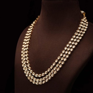 May include: A three-strand gold necklace with clear, oval-shaped stones. The necklace is displayed on a brown mannequin against a black background. The stones are set in a gold metal, creating a luxurious look.