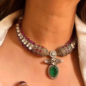 May include: An ornate necklace with a curved design, featuring alternating ruby and clear gemstone accents. The pendant has a large, oval emerald surrounded by smaller stones. A statement piece of jewellery.