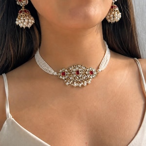 Puede incluir: Un collar tipo gargantilla dorado con un colgante central de piedras cuadradas rojas y cristales transparentes, acentuado con pequeñas perlas blancas. Se acompaña de pendientes jhumka a juego en forma de campana con piedras rojas y perlas blancas.