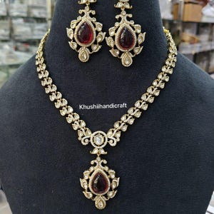 Peut inclure: Un ensemble collier et boucles d'oreilles de couleur dorée. Le collier présente un pendentif central avec une grande pierre rouge foncé en forme de goutte, entourée de petites pierres transparentes. Les boucles d'oreilles assorties ont également des pierres rouges en forme de goutte.