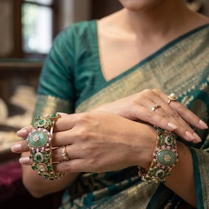 May include: A pair of gold-toned metal bangles featuring large central oval green stones, surrounded by smaller green and pink accent stones, and clear crystals. The bangles are worn on the wrists, complemented by three gold-toned rings with small stones.