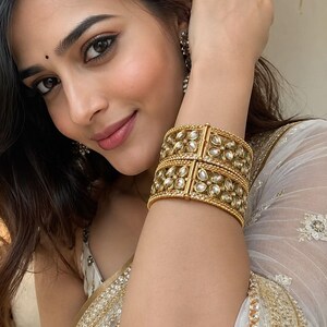 May include: A pair of wide gold-toned metal bangles featuring a repeating floral pattern inlaid with clear, teardrop-shaped stones. The bracelets have a textured, beaded border along the top and bottom edges and a hinged clasp mechanism for closure.