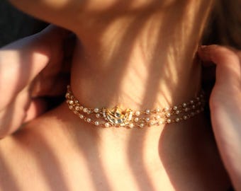 Puede incluir: Un collar gargantilla dorado con tres hilos de pequeñas perlas. El collar presenta un colgante central con un diseño decorativo y pequeñas piedras preciosas. La imagen está iluminada por la luz del sol, creando sombras en la piel.