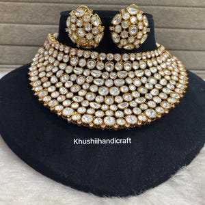 Op de afbeelding: Een goudkleurige ketting en bijpassende oorbellen set. De sieraden hebben talrijke heldere, ronde edelstenen die dicht op elkaar zijn gezet. De ketting is een choker-stijl en de oorbellen zijn rond. De set wordt gepresenteerd op een zwart fluwelen oppervlak.