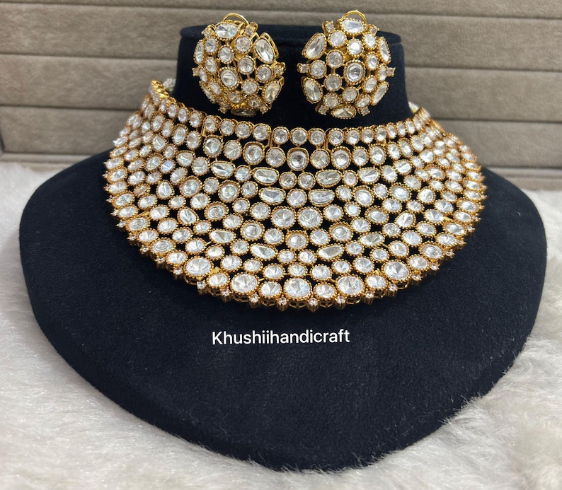 Op de afbeelding: Een goudkleurige ketting en bijpassende oorbellen set. De sieraden hebben talrijke heldere, ronde edelstenen die dicht op elkaar zijn gezet. De ketting is een choker-stijl en de oorbellen zijn rond. De set wordt gepresenteerd op een zwart fluwelen oppervlak.