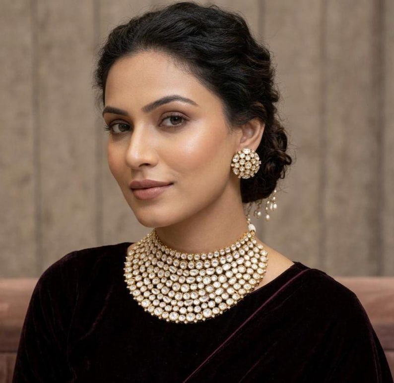Op de afbeelding: Een vrouw draagt een goudkleurige ketting en bijpassende oorbellen. De ketting is een meerlagige choker-stijl en de oorbellen zijn ronde studs. De sieraden zijn versierd met heldere, sprankelende stenen. De vrouw draagt een donker bordeauxrode top.
