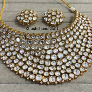 Op de afbeelding: Een goudkleurige ketting en bijpassende oorbellen set. De sieraden hebben talrijke heldere, ronde en ovale stenen die dicht op elkaar zijn geplaatst. De tekst "Khushiihandicraft" is zichtbaar op de afbeelding.