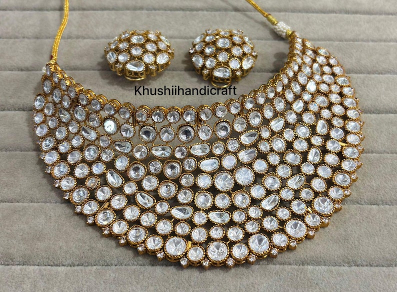 Op de afbeelding: Een goudkleurige ketting en bijpassende oorbellen set. De sieraden hebben talrijke heldere, ronde en ovale stenen die dicht op elkaar zijn geplaatst. De tekst "Khushiihandicraft" is zichtbaar op de afbeelding.