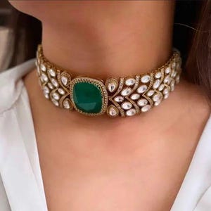 Könnte beinhalten: Ein goldfarbenes Choker-Halsband mit einem großen, quadratischen, smaragdgrünen Edelstein in der Mitte. Die Halskette ist mit zahlreichen klaren, funkelnden Edelsteinen und aufwendigen Details verziert, was ein luxuriöses und elegantes Aussehen erzeugt.