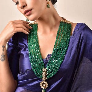 Puede incluir: Collar de cuentas verde esmeralda de múltiples hilos con un colgante dorado con un diseño floral y una pequeña piedra preciosa verde. También se ven pendientes verdes esmeralda a juego. El collar se lleva sobre una blusa de satén azul.