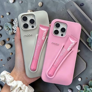 May include: Two phone cases, one light gray and one pink, each with a matching pink lip product. The cases have the brand name "rhode" printed on them. The lip products are in pink tubes with white text.