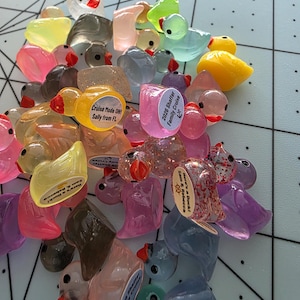 May include: A collection of colorful, translucent duck figurines in various shades of pink, yellow, blue, and green. Some ducks have small labels with text. The ducks are scattered on a light gray surface.