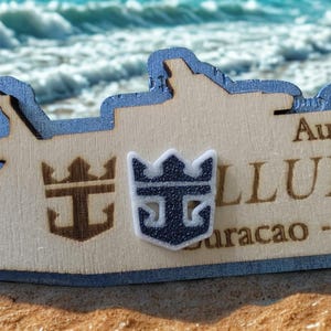 Könnte beinhalten: Eine lasergeschnittene Kreuzfahrtschiff-Silhouette aus Holz mit blau lackiertem Rand, einem dunkelbraun gravierten Kronenlogo, einem erhabenen weiß-dunkelblauen Kronenemblem und eingraviertem Text August 2024, ALLURE OF THE SEAS, Curacao - Aruba.