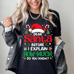 May include: Black t-shirt with a Christmas graphic. The design features a red Santa hat, candy canes, and the text "Dear Santa, Before I Explain, How Much Do You Know?" in red and green.