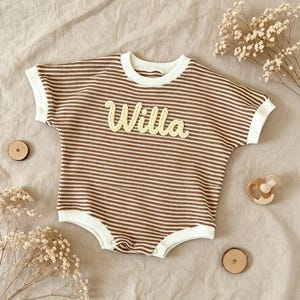May include: A brown and white striped baby romper with the name "Willa" embroidered in yellow script. The romper has short sleeves and white trim. The image includes a pacifier, wooden discs, and dried flowers.