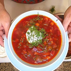 Peut inclure: Un bol de bortsch rouge vif, garni d'une cuillerée de crème blanche et d'aneth frais. La soupe contient des légumes visibles. Le bol est tenu par des mains, suggérant un repas fait maison.