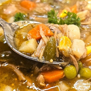 Pode incluir: Uma tigela de sopa com uma colher cheia de vegetais e carne. A sopa contém cenouras, ervilhas, milho e batatas. A sopa é de cor castanha escura e está numa tigela branca.