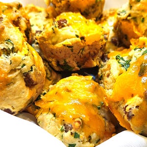 May include: A basket filled with freshly baked muffins. The muffins are golden brown with melted orange cheese on top. Green herbs are visible throughout the muffins. The muffins are in a white basket with a black rim.