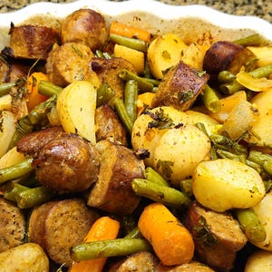 May include: A close-up shot of a baked dish filled with roasted sausage, potatoes, carrots, and green beans. The ingredients are arranged in a white, scalloped-edge dish. The food is seasoned with herbs and spices, and the colors are warm and inviting.
