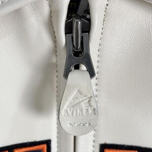 May include: Close-up of a white leather jacket zipper. The zipper is silver-toned with the brand name "AVIREX" engraved on the pull tab and the zipper itself. The jacket has white stitching and a textured surface.
