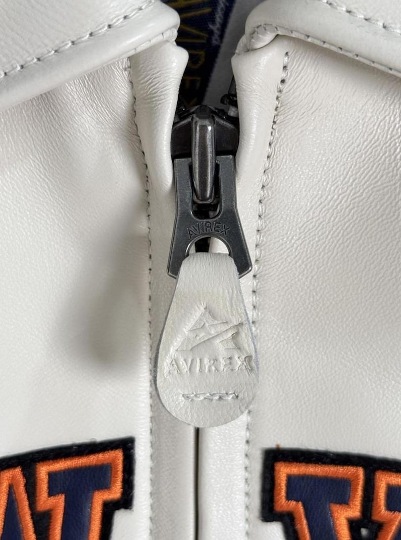 May include: Close-up of a white leather jacket zipper. The zipper is silver-toned with the brand name "AVIREX" engraved on the pull tab and the zipper itself. The jacket has white stitching and a textured surface.