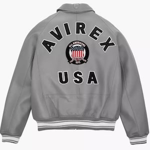 May include: A gray leather jacket with the word "AVIREX" arched across the upper back in black letters. Below, the word "USA" is displayed in black block letters. A circular patch with the words "ALL AMERICANS" and "ESTABLISHED 1975" surrounds a red, white, and blue design.
