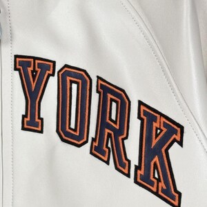 May include: Close-up of a white leather jacket with the word "YORK" embroidered on the front. The letters are navy blue with orange outlines. The jacket has a collar and a zipper.